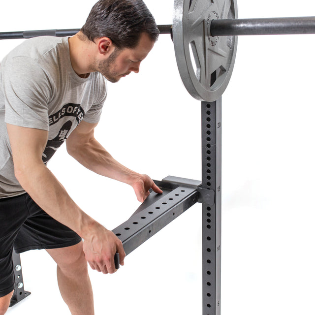 A man in a gray t-shirt and black shorts adjusts a squat rack's safety bar loaded with weight plates, aligning it precisely with the Bells of Steel Spotter Arms Rack Attachment. The plain white background highlights his meticulous preparation for barbell supersets.