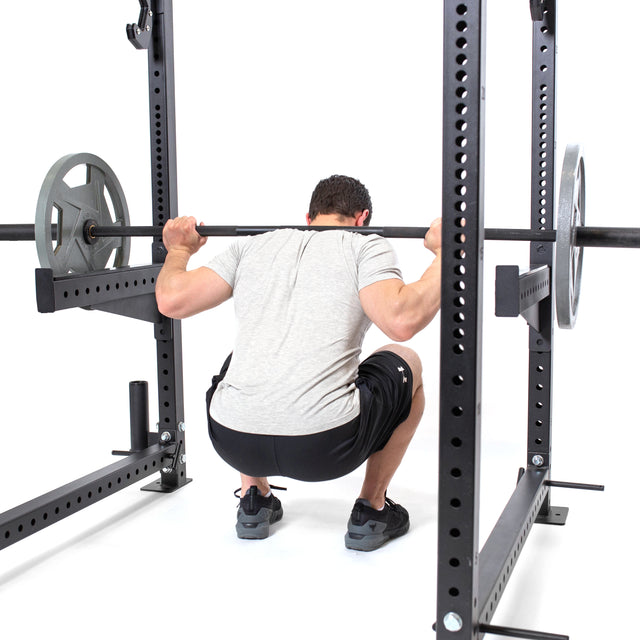 Wearing a gray shirt and black shorts, a person squats with a barbell on their back in a rack equipped with Bells of Steel's Spotter Arms Rack Attachment for safety, while UHMW lining protects the frame and weight plates are loaded.