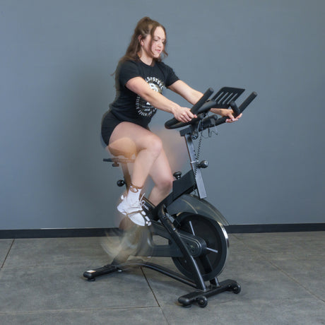A woman in athletic wear rides the Bells of Steel Blitz Indoor Cycling Bike indoors against a plain gray wall, her legs blurred in motion as she pedals on the adjustable exercise bike.