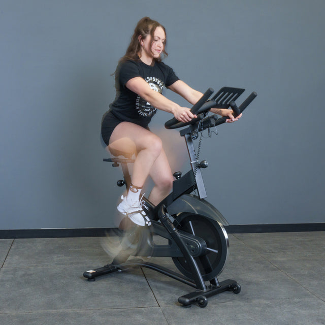 A woman in athletic wear rides the Bells of Steel Blitz Indoor Cycling Bike indoors against a plain gray wall, her legs blurred in motion as she pedals on the adjustable exercise bike.