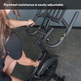 A person adjusts the resistance knob on the Bells of Steel Blitz Indoor Cycling Bike, with its display and handlebars visible. Text at the top reads, "Easily adjust flywheel resistance on this magnetic indoor cycling bike.
