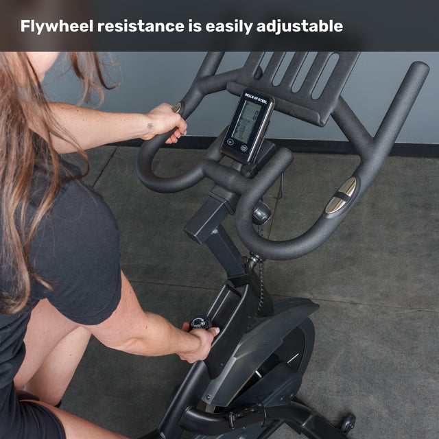 A person adjusts the resistance knob on the Bells of Steel Blitz Indoor Cycling Bike, with its display and handlebars visible. Text at the top reads, "Easily adjust flywheel resistance on this magnetic indoor cycling bike.
