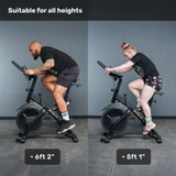 Two riders, 6'2" and 5'1", use the Bells of Steel Blitz Indoor Cycling Bike side by side. Text above says "Suitable for all heights." Both are comfortably positioned thanks to the bike’s adjustable features.