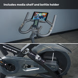 Close-up of the Bells of Steel Blitz Indoor Cycling Bike displaying its media shelf with tablet, water bottle holder, pedals, and flywheel, plus text above: "Includes media shelf and bottle holder.