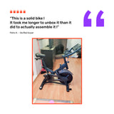 A Bells of Steel Blitz Indoor Cycling Bike stands on a wooden living room floor by a fireplace, with a review from verified buyer Petro K above, highlighting the bike’s easy assembly.