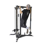 A person in a black shirt, gray pants, cap, and sneakers performs a pull-up using the Bells of Steel All-in-One Trainer with various pulleys and weights on a white background.