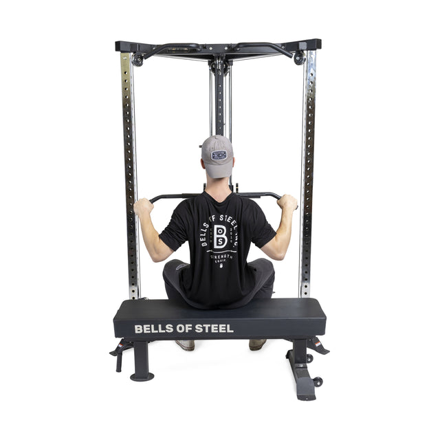 A person wearing a black shirt and cap uses Bells of Steel's All-in-One Trainer; seated, they pull down on the lat pulldown bar, highlighting the versatile design and features of this multifunctional equipment.