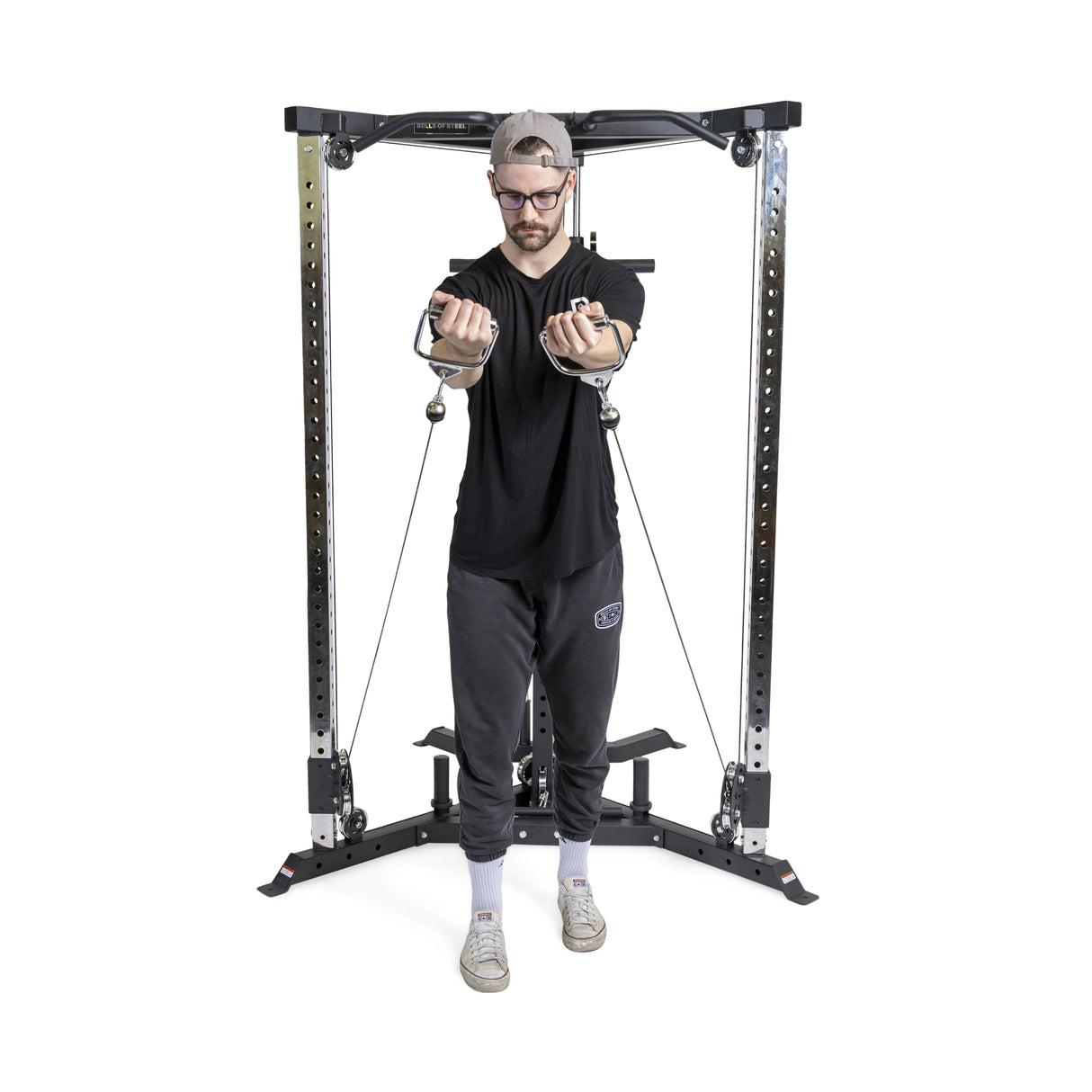 Exercising on a Bells of Steel All-in-One Trainer, a person, in a black shirt, dark pants, cap, and glasses, pulls handles towards their chest against a white backdrop.