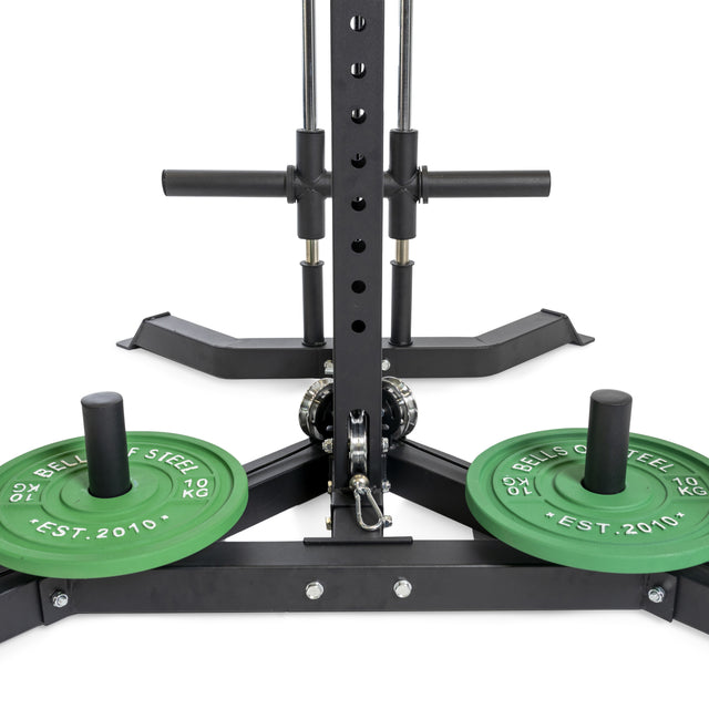 Close-up of Bells of Steel's All-in-One Trainer, a versatile power rack doubling as a squat stand. It includes two 10 kg green plates marked "Bells of Steel" at the base and features adjustable metal bars and pegs for all your fitness needs.