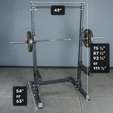 A Bells of Steel Manticore Squat Stand Builder (3" x 3", 1" Holes) in black, featuring 3x3 uprights and a barbell, with labeled dimensions: width of 49", height options of 75¼", 87¼", 93¼", or 111¼", and base height choices of either 54" or 63". Perfect for home gym setups, the squat stand is positioned on a gray tiled floor against a plain wall background.