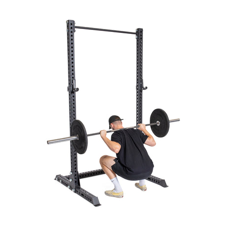 Someone in athletic wear squats with a barbell on their shoulders using a Bells of Steel Manticore Squat Stand Builder, featuring 1" adjustment holes. The sturdy setup highlights its 1,000-pound capacity, ensuring stability and safety during intense workouts.