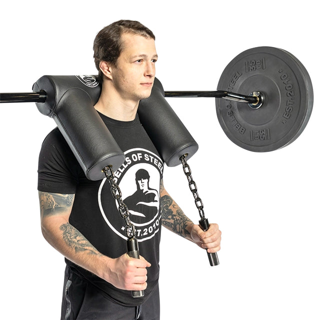 A person is squatting with Bells of Steel's Safety Squat Bar - The SS4, using its ergonomic pad for support. They're wearing a black t-shirt featuring a logo and displaying tattoos on their arms.