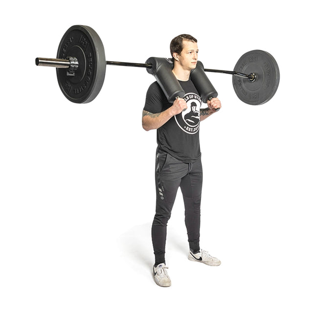 A person stands with the Bells of Steel Safety Squat Bar - The SS4 on their shoulders, wearing a black T-shirt and dark pants. The weights are supported by an ergonomically designed pad, set against a plain white background.