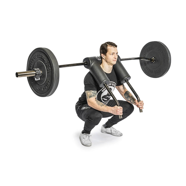 Wearing a black shirt, pants, and gray shoes, a person performs squats with Bells of Steel's Safety Squat Bar - The SS4. The ergonomic pad ensures comfort while they hold a low squat position, the barbell securely resting on their shoulders.