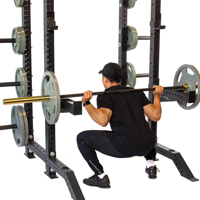 A person wearing black workout clothes and a cap is performing a squat in the gym, expertly using barbell supersets. They're utilizing the Bells of Steel Spotter Arms Rack Attachment for added safety as they manage the weights with precision.