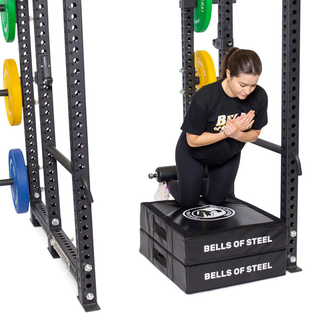 A woman does a Nordic hamstring curl in the gym using a Split Squat Leg Roller Rack Attachment. Kneeling on a padded surface labeled "Bells of Steel," she positions herself within a squat rack with colored weights, hands together.