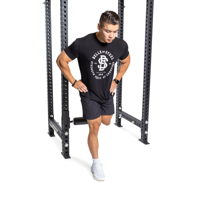 A man in a black T-shirt and shorts stands in a power rack, ready to exercise with the Bells of Steel Split Squat Leg Roller Rack Attachment. He leans slightly forward, hands on hips, wearing white shoes. The background is plain white.