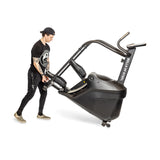 A person tilts and moves the Bells of Steel Blitz Stepper—a large curved treadmill with handles and wheels—on a white background, designed for low-impact cardio workouts.