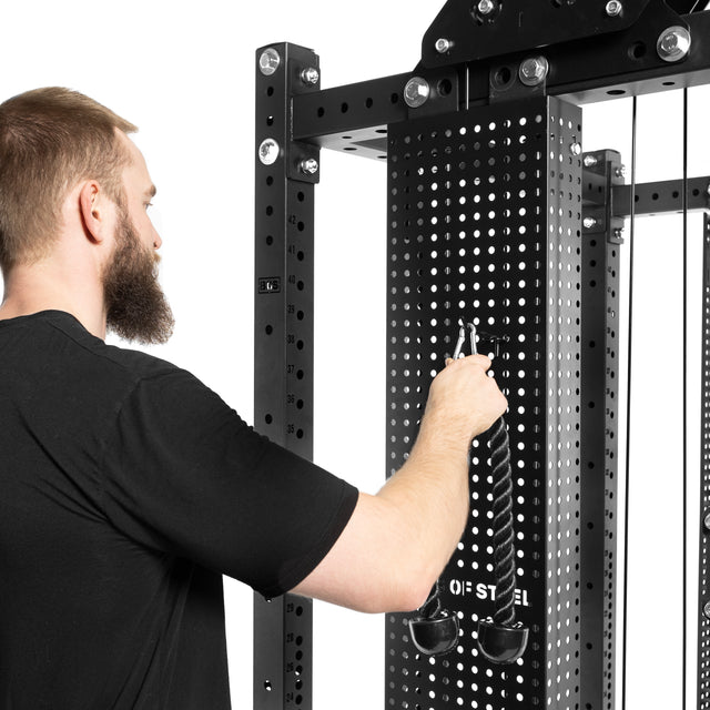 A bearded man in a black shirt is attaching a rope handle to a Kraken Weight Stack Shroud by Bells of Steel in the gym. This sleek home gym equipment features metallic components and perforated panels.