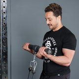 A man in a black Bells of Steel t-shirt does a cable bicep curl in the gym, using the Bells of Steel Fat Bar – Straight Handle Bar attached to resistance cables while looking down at his arms.