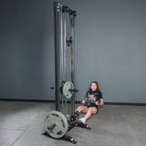 A woman prepares for a seated row in a gym, holding the Bells of Steel Fat Bar - Straight Handle Bar, which is attached to a cable machine with weight plates. The gym features gray walls and a tiled floor.