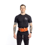 A person stands against a white background, donning a black T-shirt with "BOS" and black pants. Tattoos adorn both arms, while a Bells of Steel USA 10mm Leather Weightlifting Belt cinches their waist, adding an extra edge to their stylish ensemble.