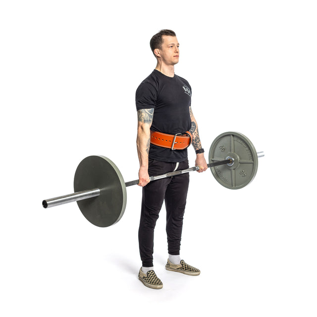 A person stands holding a barbell, wearing a black t-shirt, black pants, checkered shoes, and showing tattoos on their left arm. They wear a 10mm Leather Weightlifting Belt from Bells of Steel USA. The background is white.