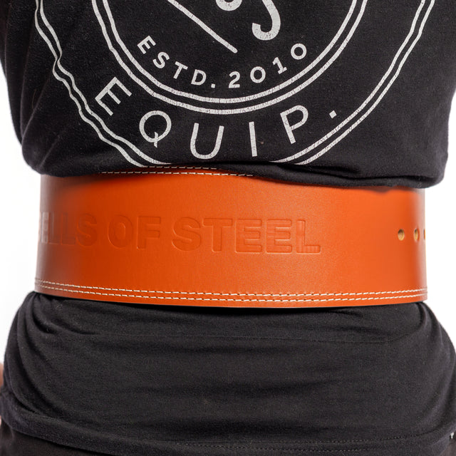 A person wearing a black shirt with a circular logo dons an orange 10mm Leather Weightlifting Belt by Bells of Steel USA, featuring "BELLS OF STEEL" embossing and a single-prong design for secure support.