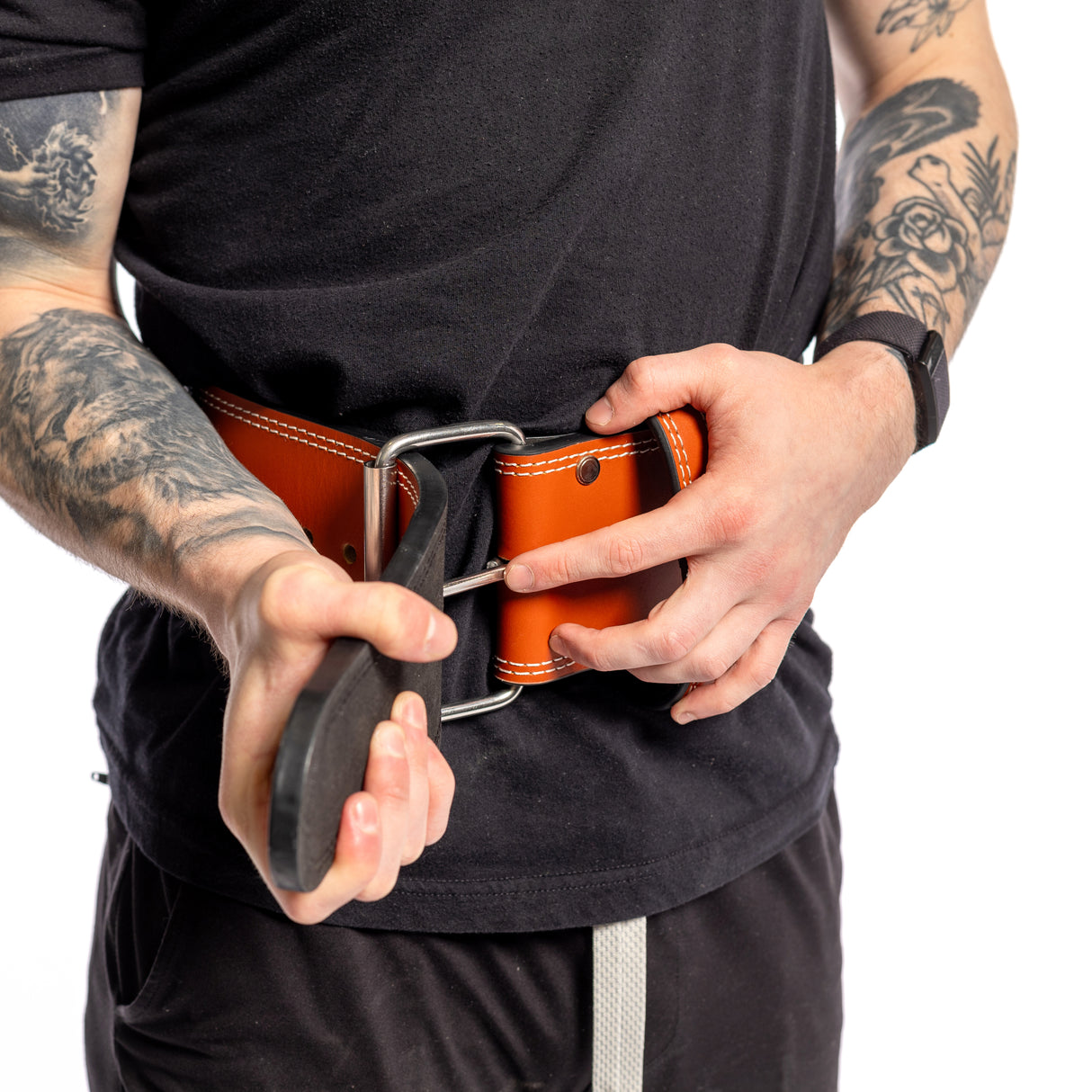 A tattooed person, wearing a black t-shirt, adjusts a Bells of Steel USA 10mm Leather Weightlifting Belt around their waist, gripping the strap with both hands. The single-prong belt boasts sturdy metal buckles.