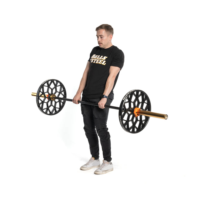 A man in a black "Bells of Steel" shirt and black jeans lifts a barbell with Technique Plates - 5 Lbs (Pair), coated in virgin rubber. He is standing against a plain white background, looking focused.