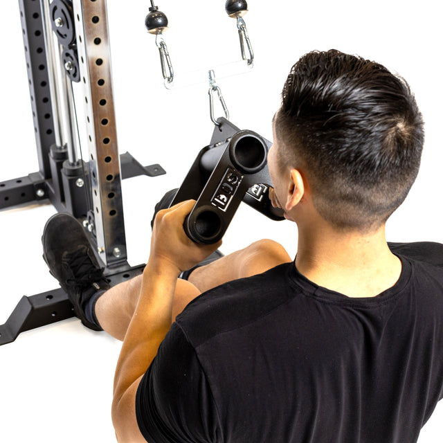 A person with short dark hair is seated on the floor, performing a cable row exercise using the Bells of Steel Fat Bar - T-Bar Row Attachment to enhance grip strength. They're wearing a black shirt and shorts, holding the double grip handle, and facing away from the camera.