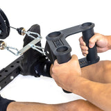 A person holds the Bells of Steel Fat Bar - T-Bar Row Cable Attachment, ready for their workout. The black machine, with weights and metal cables, enhances grip strength. Their slightly bent legs in black shoes emphasize the focus on this equipment's handle.