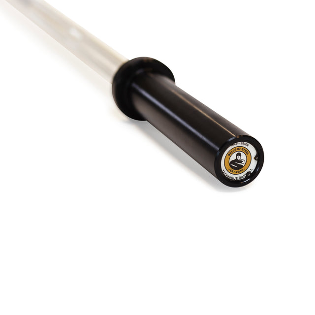 A close-up of a Bells of Steel Aluminum Technique Barbell end with a black sleeve and branding label. The metallic finish showcases the weightlifting figure logo. Perfect for mastering barbell lifts, with the bar extending into a slightly blurred background.