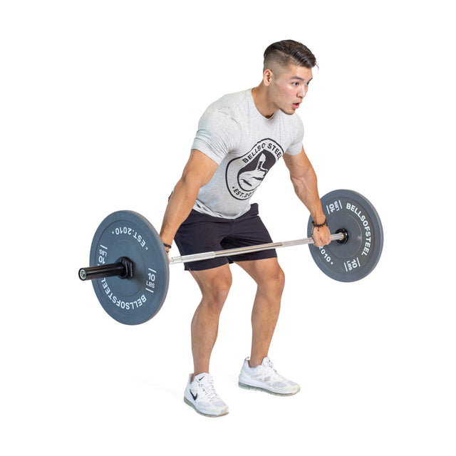 A focused individual in a gray T-shirt, black shorts, and white sneakers is lifting the Bells of Steel Aluminum Technique Barbell on a white-background gym set, perfecting their deadlift form.