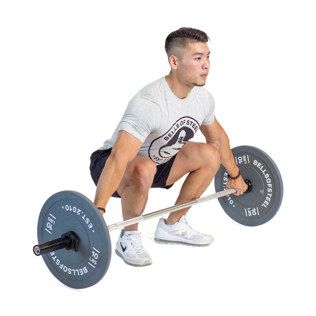 A person wearing a gray T-shirt and black shorts performs a deadlift using a Bells of Steel Aluminum Technique Barbell. They are squatting, holding the bar with both hands, focused on their exercise.