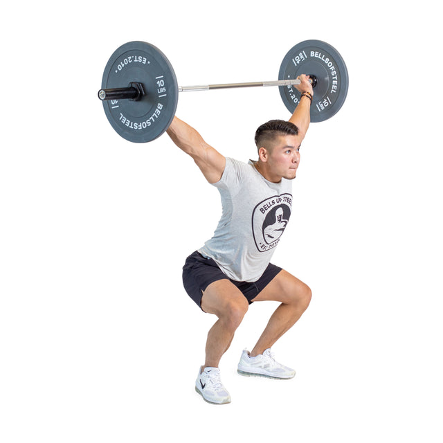 A person mastering an overhead squat with a Bells of Steel Aluminum Technique Barbell, dressed in a gray t-shirt and black shorts. The plain white background highlights their lifting skills and exercise form.