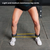 A person stands with legs apart, wearing white shoes and black shorts. Yellow resistance bands from the Bells of Steel USA Agility Ladder and Leg Cord Bundle are strapped between their ankles on a gym floor, perfect for effective agility training.