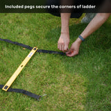 A trainer secures the yellow Bells of Steel USA Agility Ladder and Leg Cord Bundle to the grass with black straps and included pegs. Text reads, “Included pegs secure the corners of ladder—perfect for your agility training or speed and agility kit.”.