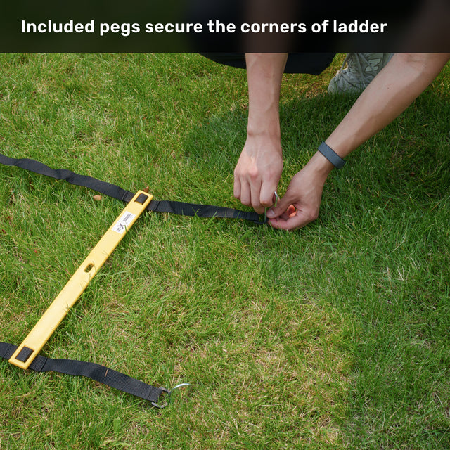 A trainer secures the yellow Bells of Steel USA Agility Ladder and Leg Cord Bundle to the grass with black straps and included pegs. Text reads, “Included pegs secure the corners of ladder—perfect for your agility training or speed and agility kit.”.