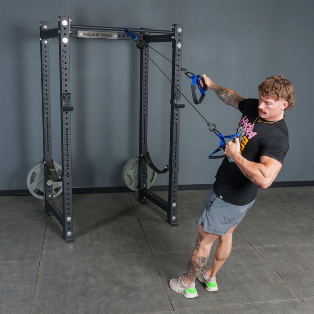 A person uses the Bells of Steel USA Rotational Pulley for rotational training, pulling on handles with one arm while leaning back, in a gym with grey walls and flooring.