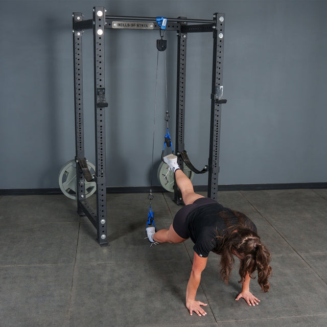 Using the Rotational Pulley from Bells of Steel USA, a person performs an intense bodyweight workout by suspending their feet in straps attached to a squat rack and balancing on their hands on the gym floor.