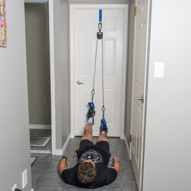 A person lies on the floor with their feet strapped into the Bells of Steel USA Rotational Pulley, using resistance bands for a leg workout or physical therapy exercise that can easily be incorporated into a bodyweight workout routine.