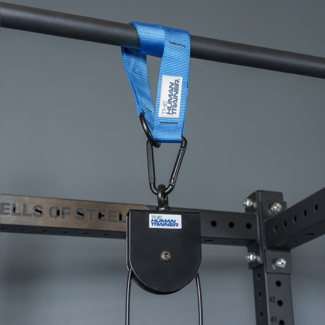 A blue strap labeled “The Human Trainer” loops over a pull-up bar, attached to the Bells of Steel USA Rotational Pulley kit designed for bodyweight workouts. In the background, a power rack with “BELLS OF STEEL” embossed on the metal frame is visible.