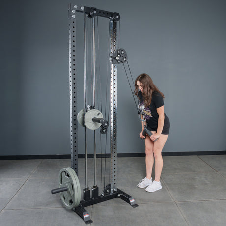 A woman in shorts and a T-shirt stands by a Bells of Steel Fat Bar - Tricep Pressdown machine in the gym, gripping the handle with both hands as she prepares for tricep pushdowns. The machine is loaded with multiple weight plates.