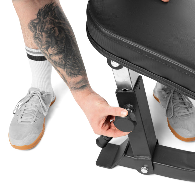 A person with a lion calf tattoo adjusts the height of the Bells of Steel Adjustable Utility Bench using a knob. Wearing white socks and gray shoes with gum soles, they use the 575lb max capacity bench for versatile workouts.