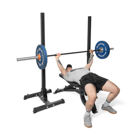 A person in athletic wear performs a bench press on the Bells of Steel Adjustable Utility Bench, lifting a barbell with blue plates inside a black power rack with a 575lb max capacity.