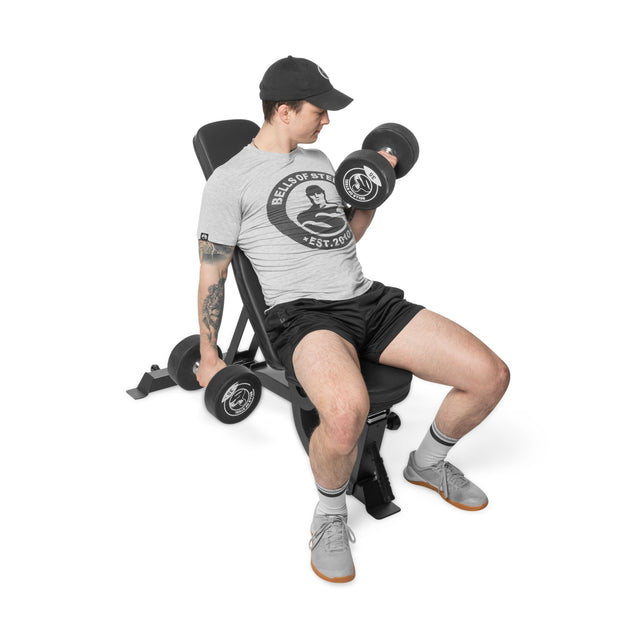 A man in athletic wear and a cap sits on the Bells of Steel Adjustable Utility Bench, performing seated dumbbell curls with both arms against a plain white background.
