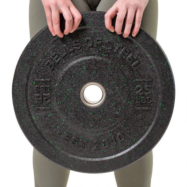 A person in green leggings holds a large Crumb Bumper Plate from Bells of Steel with green speckles, confidently gripping the 25 lbs weight, showcasing their dedication to lifting.