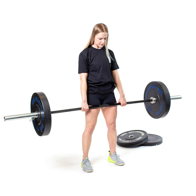 A person lifts the Bells of Steel Multi-Purpose Olympic Barbell – The Utility Bar in a gym. They're dressed in a black T-shirt, black shorts, and bright sneakers, with more weights stacked on the floor nearby.