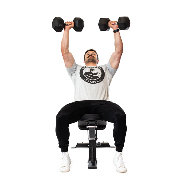 A man lifts two dumbbells overhead while seated on a Bells of Steel Adjustable Utility Bench. Wearing a gray T-shirt, black pants, and white sneakers, he seamlessly adjusts through different incline angles. The background is plain white.